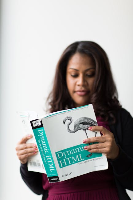 A woman with long dark hair, dressed in a black blazer and maroon top, is holding an open booklet titled 'Dynamic HTML' with a flamingo illustration on the cover. She is indoors against a plain white
