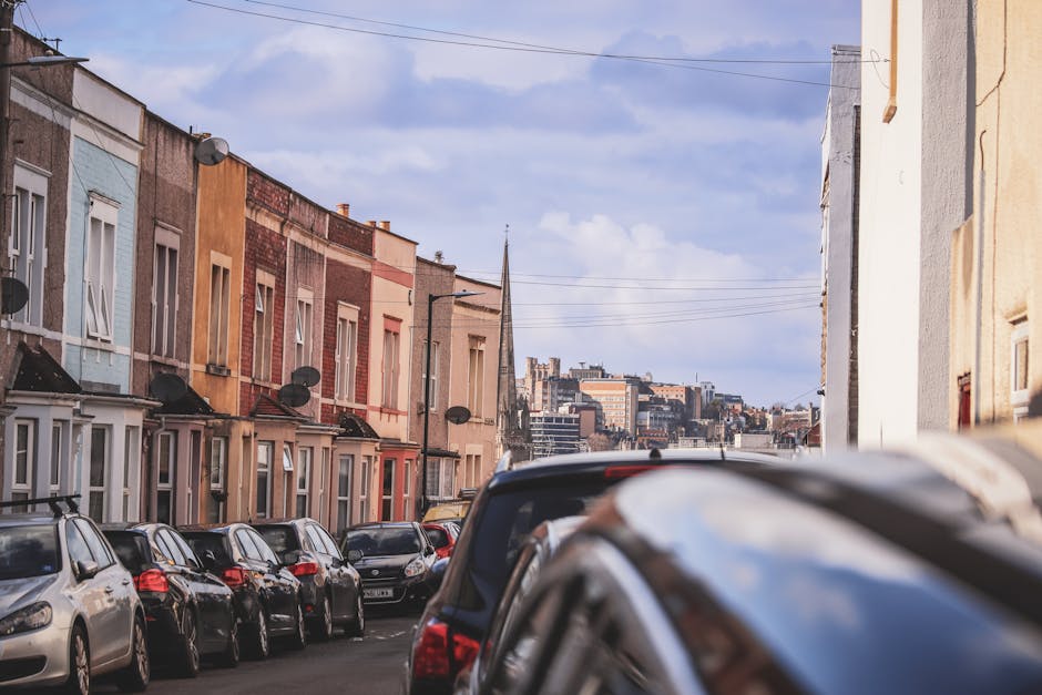 A narrow residential street in Wales lined with terraced houses painted in various pastel shades, with small front gardens, some with fences or hedges. Parked cars occupy both sides of the street, inc